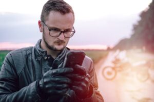 A young man in glasses and leather jacket uses a smartphone with a motorcycle in the background, symbolizing assistance and connectivity while traveling on the road.