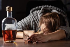 A woman who is drunk and intoxicated is sleeping near a glass of whiskey. She is a heavy drinker and alcoholic, passed out and lying asleep after drinking, highlighting issues related to alcoholism. 