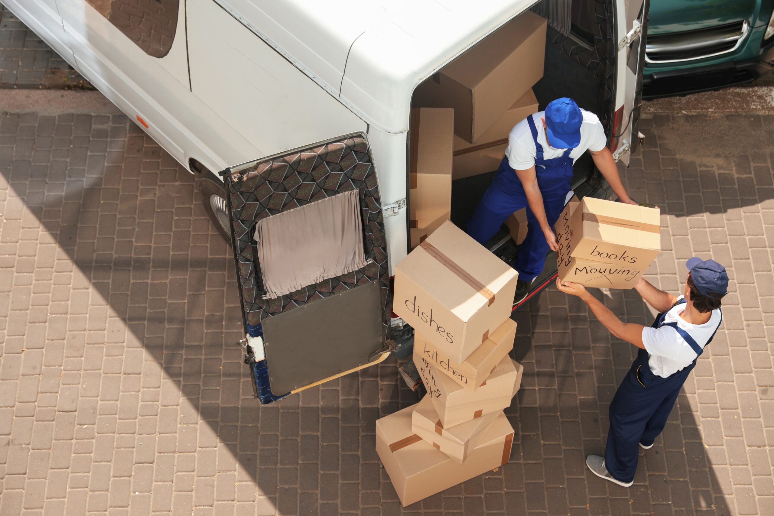 op-down view of male movers unloading boxes from a van outdoors. The movers are wearing casual clothing and are in the process of lifting or carrying cardboard boxes. The van is parked on a paved surface, and there is green grass and a building visible in the background.