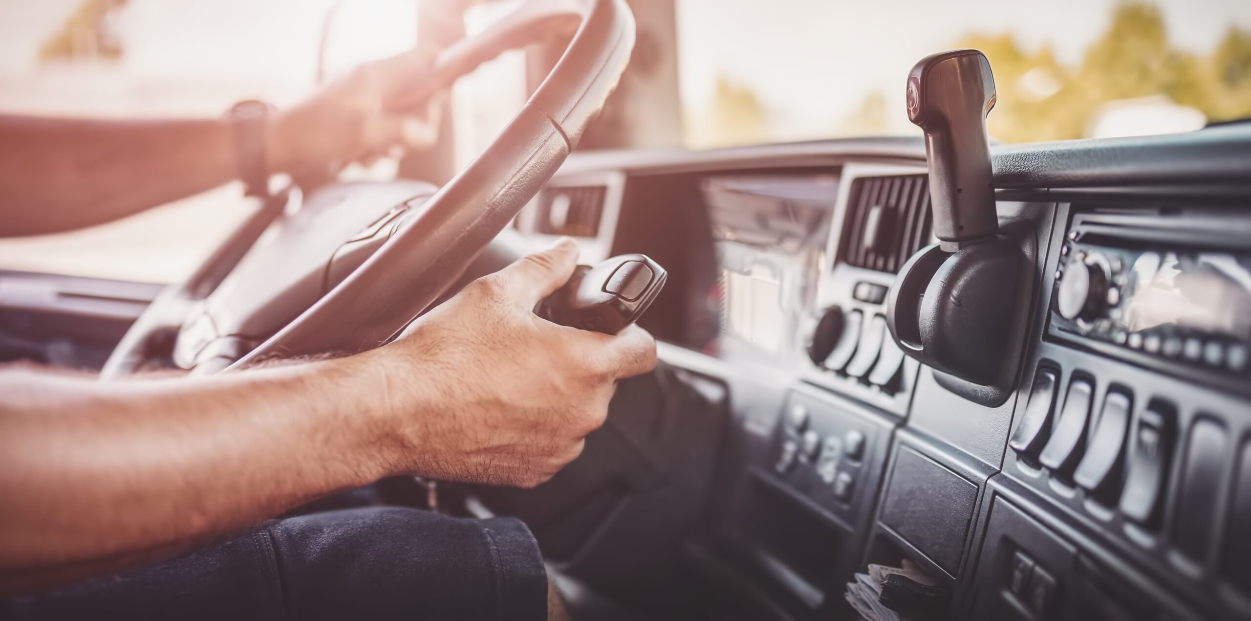 A driver sitting in the cab of a semi-truck, holding the steering wheel while driving. The driver is focused on the road ahead, wearing casual clothing, and the interior of the truck is visible, with various controls and the windshield showing a view of the road outside.