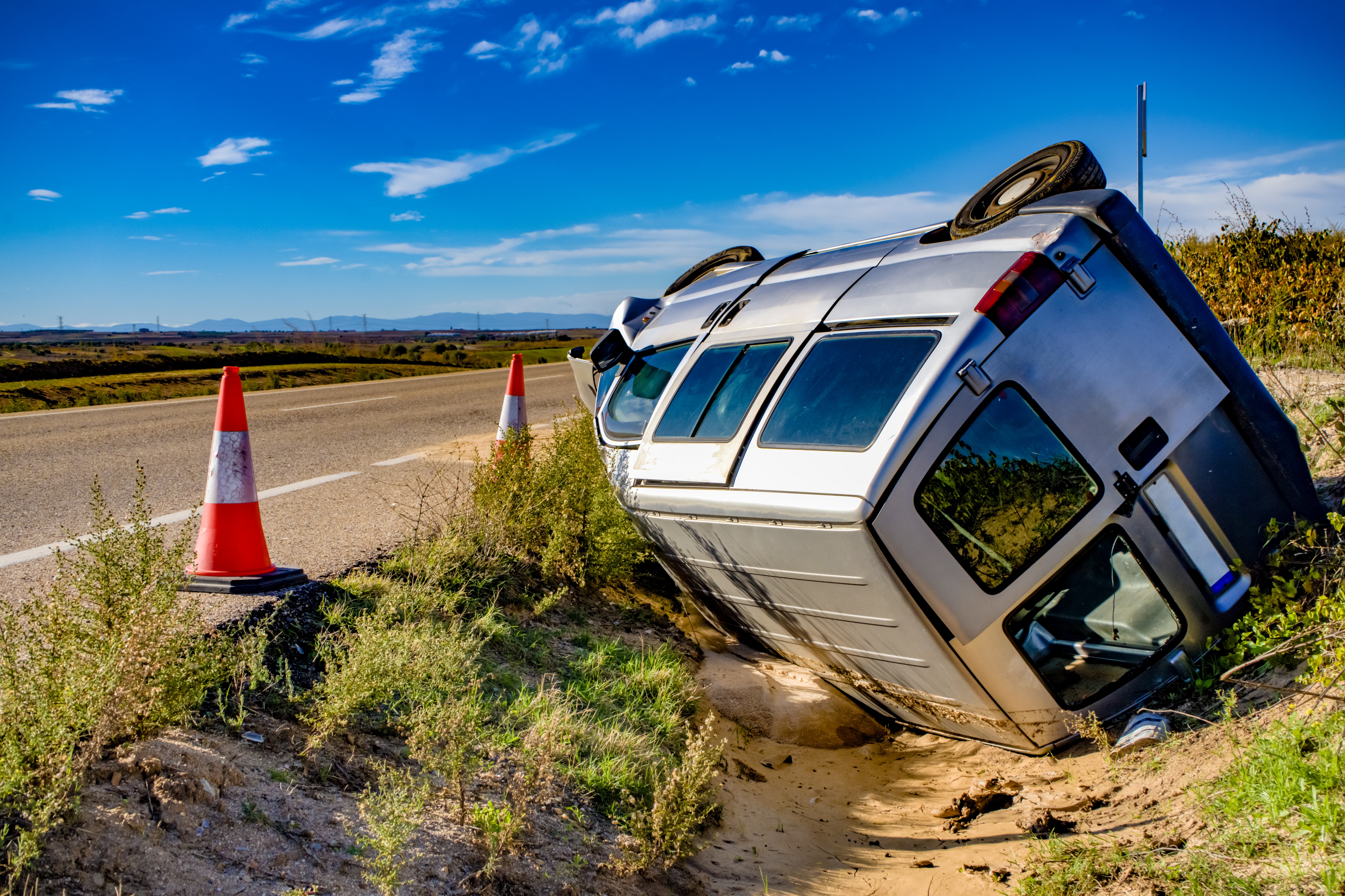 An old, damaged van is parked on the side of a road. The van shows visible signs of wear, with dents and broken windows. The surrounding area features asphalt, grass, and trees, and the scene is set in an outdoor, rural or suburban environment