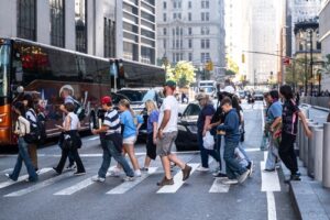 A group of pedestrians crosses a busy street in a city, surrounded by tall buildings, cars, and a parked bus in the background.