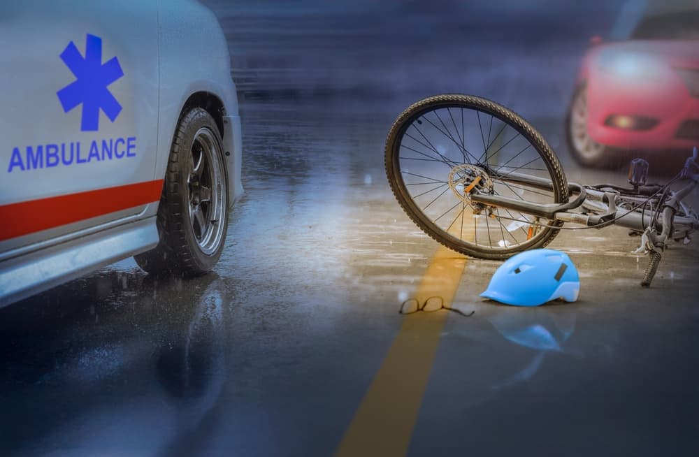 Bicycle accident scene showing a fallen bike, helmet, and eyeglasses on a wet road beside an ambulance after a vehicle collision.