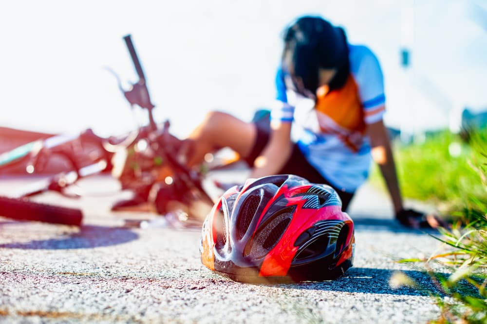 Cyclist sitting injured on the ground after a crash with a helmet in the foreground, illustrating the serious risk of road rash and deep skin abrasions.