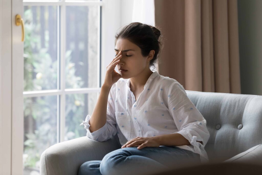 Stressed woman sitting on couch at home with hand on face, experiencing emotional distress or fatigue.